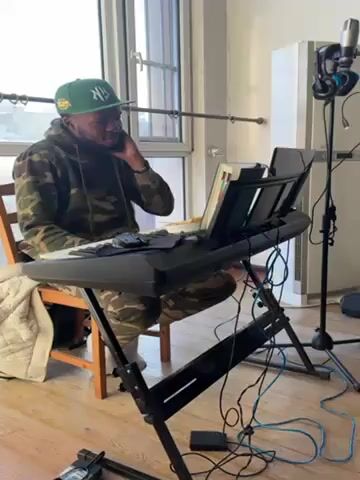 A man is sitting at a keyboard, playing it. He wears a green baseball cap with a yellow logo, a camo jacket, and camo pants.  A stand holds sheet music on the keyboard.  Various wires run from the keyboard to the floor.  To the right of the keyboard, a microphone and headphones are mounted on a stand.  Behind that is a white air conditioner.  The room is white with light wood floors.  The man looks down at the keyboard.  A chair and a blanket are to his left.