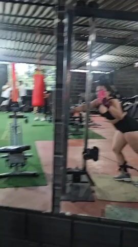 The video shows the interior of a gym, viewed through a window. The gym has a green, artificial turf floor, exposed metal beams in the ceiling, and black brick accent walls. Gym equipment, including weight benches, a squat rack, dumbbells, and a punching bag, are seen throughout. There are a handful of people working out. A woman in a black sports bra and shorts stands in front of a cable machine, performing a leg workout. She wears ankle straps connected to the cable and holds her phone up, presumably taking a picture or video.
