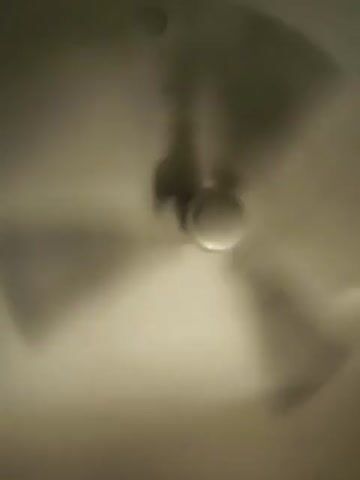 The video shows a ceiling fan in motion. The fan has four blades and a central motor housing. The blades are blurred due to their speed, creating a halo effect around the central housing. Shadows cast by the fan blades are visible on the ceiling, also blurred. The lighting is dim, creating a muted, brownish-yellow hue across the scene. The camera appears to be positioned directly below the fan, looking upwards. The overall impression is one of rapid movement and a slightly obscured view due to the motion blur.