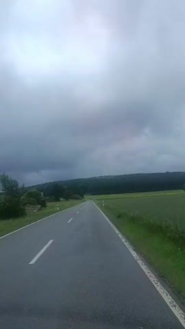 The video shows a road in a rural area. The road is paved and has white lines marking the lanes. The road is surrounded by green fields and trees. The sky is cloudy and appears to be overcast. The video is shot from a moving vehicle, as indicated by the slight blurring of the background and the perspective of the road. There are no people or animals visible in the video. The overall impression is one of a calm, ordinary drive through the countryside.