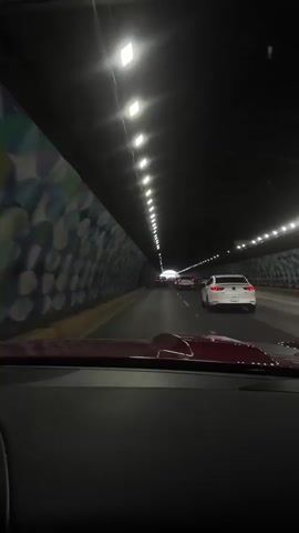 The video shows a point-of-view shot from inside a car driving through a tunnel. The tunnel has a dark ceiling with a series of evenly spaced lights. The walls of the tunnel are decorated with a colorful, abstract mosaic pattern. There are several cars in front of the camera car, also driving through the tunnel. The traffic appears to be moderate, with cars spaced a reasonable distance apart. The overall lighting in the tunnel is dim, but the overhead lights illuminate the roadway. The interior of the car is mostly dark, with only the dashboard and the hood of the car partially visible in the foreground.