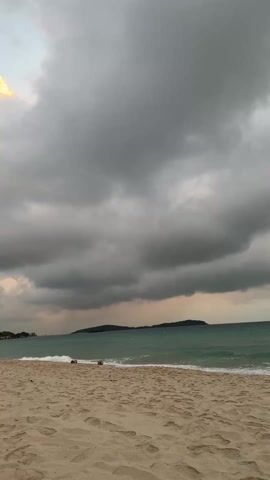 The video shows a beach under a cloudy sky. The sand is light-colored and patterned with footprints. The water is a greenish-blue with small waves gently rolling in. In the distance, there are some dark shapes that could be islands or land formations. As the video pans, more land mass is visible. There appears to be a small figure on the sand towards the left in the beginning of the video. The sky is filled with dark, ominous clouds, though there is some light peeking through near the horizon.