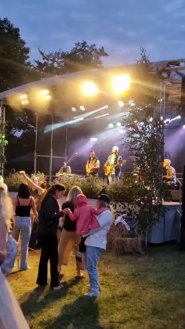The video shows an outdoor evening concert. A band is performing on a raised stage, lit by bright stage lights and purple uplighting. There are several band members on stage, some playing guitars. In front of the stage, a crowd of people are gathered. In the foreground, a couple is dancing. A woman is holding a small child. The environment appears to be a grassy area with trees in the background. There are hay bales near the stage, possibly for seating or decoration. The sky is dark, indicating it is evening.