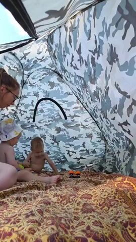 Inside a camouflage-patterned tent, a woman with glasses and her hair in a ponytail sits on a patterned blanket. She is facing two young children. To her left, a baby in a sun hat sits on the blanket. To her right, a toddler in red shorts is crawling and playing with a small orange toy car. The tent interior is visible, along with a glimpse of the sky outside.