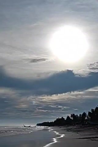This video shows a beach with a body of water, sand, and trees. The sun is visible in the sky, and clouds are present. There is a boat in the water near the shore. The waves are gently lapping on the beach, and the sand is wet from the tide. The trees are silhouetted against the sky, and their leaves are rustling in the breeze. The overall atmosphere is peaceful and serene.