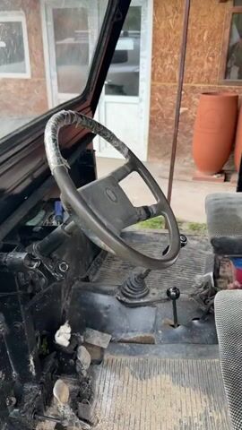 The video shows a black, custom-built vehicle that appears to be undergoing repair. The interior of the cab is visible, showing the steering wheel, pedals, and gear shift. The front hood is partially open, revealing the engine, which has a yellow component. The vehicle is parked outside, near a building with a wooden exterior and a white-framed window. The ground around the vehicle is covered with grass and dirt. The video presents a close-up view of the vehicle, highlighting its unique construction and the ongoing mechanical work.