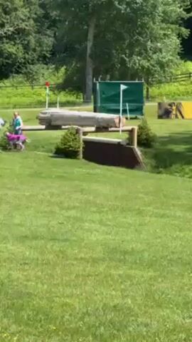 Here is a description of the video:

The video appears to show a cross-country horse riding event. The initial shot shows a grassy field with an obstacle in the middle distance. The camera pans left, following the path of the course and revealing a variety of other obstacles, including a water obstacle. A few people can be seen in the background, walking around the course. The day is sunny, with some scattered clouds in the sky.