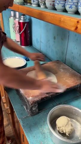 The video shows a person in a colorful patterned garment rolling out dough on a wooden board. The person is using a wooden rolling pin, and flour is dusted on the board and the dough. The dough is thin and circular. To the left of the rolling pin, there is a white plate with a piece of rolled-out dough on it. To the right of the wooden board, there is a silver bowl containing a ball of dough. The background shows a blue tiled wall with shelves above. The shelves are filled with various ceramic cups and bowls. On the wall behind the person, there are several thermoses. The counter surface is a distressed blue wood.