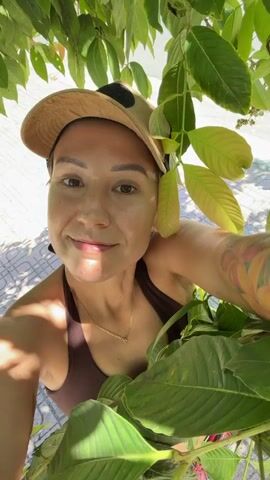 A woman wearing a brown tank top and a tan baseball cap is looking at the camera, surrounded by green leaves. She has a tattoo on her arm and is wearing a necklace. The background is blurred and appears to be outdoors. The video is short, with no discernible action.
