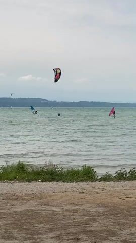 The video shows a wide shot of a body of water with several people engaged in watersports. In the foreground, there is a sandy and grassy shoreline. The water is choppy, with visible waves. In the middle ground, a kiteboarder is visible, their colorful kite high in the air. To the left, a windsurfer with a blue sail is also in the water. To the right, another windsurfer with a pink sail is visible. Further back, there are other small dark shapes in the water that could be other watersports participants or buoys. The background consists of a hazy, distant shoreline with hills or mountains and a cloudy, overcast sky. The overall impression is a day with moderate wind suitable for watersports.