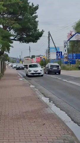 A white car is driving down a road. The car is in the right lane. There are other cars on the road as well. The road is paved and has two lanes in each direction. There is a sidewalk on the left side of the road. There are trees and buildings on the right side of the road. The sky is cloudy.
