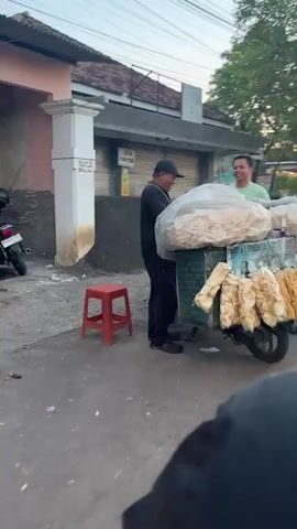 Two men are standing next to a food cart. One man, wearing a black t-shirt and a black baseball cap, is behind the cart and appears to be interacting with the other man. The second man, wearing a light green t-shirt, is standing in front of the cart, smiling. The cart is laden with large plastic bags filled with light-colored, puffy snacks, possibly crackers or chips, and several smaller bags of the same snack are hanging from the side. A red stool is placed to the left of the cart. A motorcycle is parked behind the stool. The background shows a building with closed shutters and a paved area. The lighting suggests it is either early morning or late afternoon.