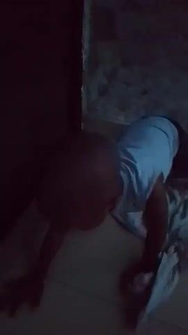 The video shows a person on all fours in a dimly lit room. The person is wearing a light-colored shirt. The environment appears to be a room with concrete walls and a tiled floor. There are some discarded clothes on the floor near the person. The camera is positioned low to the ground, looking up at the person. The person's face is not visible. The video appears to be a short clip from a longer recording.