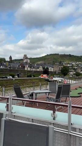 The video shows a view from a boat looking towards a town on a hillside. In the foreground, there are chairs and a table on a wooden deck, with a mini-golf course to the right. In the background, there are buildings with a church steeple, vineyards on the hillside, and a road with vehicles. The sky is cloudy.