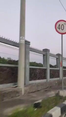 The video shows a view from a motorcycle traveling on a road. The road has a sidewalk with black and white striped curbs. On the left side of the road is a fence made of concrete pillars and stone-patterned panels. A red emblem with a yellow star is visible on one of the fence panels. There are also power lines and streetlights along the road. Several motorcycles are visible in the distance, and one person is reflected in the motorcycle's mirror, wearing a yellow jacket. The sky is overcast. A traffic sign indicating "no U-turn" is visible on a pole.