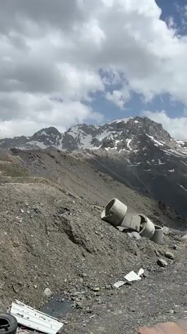 The video shows a mountainous landscape. In the foreground, there are several large concrete pipes lying on a rocky slope. Behind the pipes, a steep, gray mountain slope rises, with patches of snow visible near the peak. The mountain peak itself is rugged and covered in snow. Power lines and a utility pole are visible in the middle ground, stretching across the frame. The sky is partly cloudy, with white clouds against a blue background. The overall impression is one of a remote and rugged natural environment, possibly a construction or debris site.