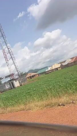 The video shows a telecommunications tower in a rural area. The tower is tall and made of metal with a white and red color scheme. At the base of the tower, there is a small white building with a cage-like structure surrounding it. The tower is situated in a field of green crops, possibly corn. In the background, there are houses with red roofs and some trees. The sky is partly cloudy with large white clouds against a blue sky. The overall impression is of an open, undeveloped landscape with essential infrastructure present.