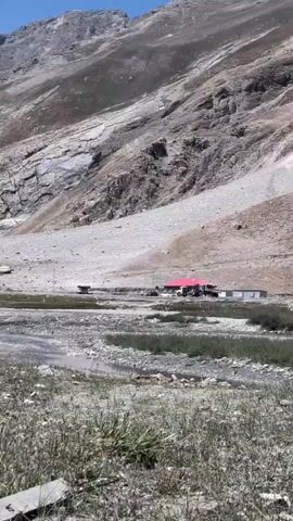 The video shows a rocky, mountainous landscape with a building with a red roof in the middle ground. Several vehicles, including what appear to be trucks and a smaller vehicle, are parked near the building and on a dirt road. The foreground is filled with dry grass and a shallow stream. The mountain slope behind the building is steep and rocky, with evidence of past rockslides or erosion. The sky is clear and bright, suggesting a sunny day.