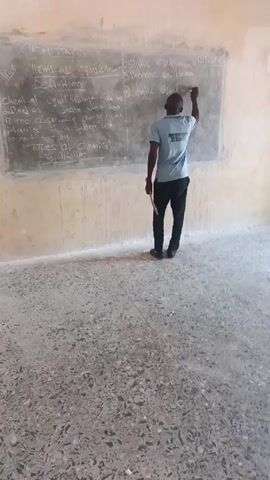 The video shows a man, from the back view and then turning to face the camera, standing in front of a chalkboard in what appears to be a classroom. The chalkboard has the heading "CHEMICAL EQUILIBRIUM" and notes written in white chalk. The man is wearing a light blue t-shirt with some writing and an emblem on the back, black pants, and black shoes. He is holding a slender wooden stick. He writes on the chalkboard, turns to look at the camera, and holds the stick casually. The background consists of a wall with a faded paint job and a tiled floor.