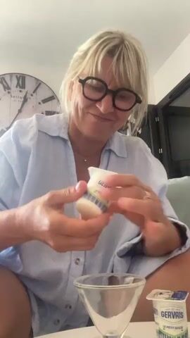 A blonde woman with glasses and a light blue shirt is sitting down and preparing to eat a serving of yogurt. She is holding a small yogurt container in her right hand and a plastic lid in her left hand. She is smiling and looking down at the yogurt. There is a glass dish in front of her, and a second yogurt container is visible on the right side of the frame. In the background, a large wall clock with Roman numerals is visible. The setting appears to be a home environment.