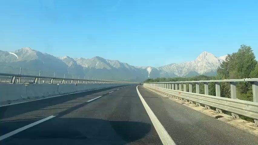 A red SUV drives on a highway with mountains in the background. The highway has two lanes, with a white line dividing them. A metal guardrail runs along the side of the road. The sky is clear and blue.