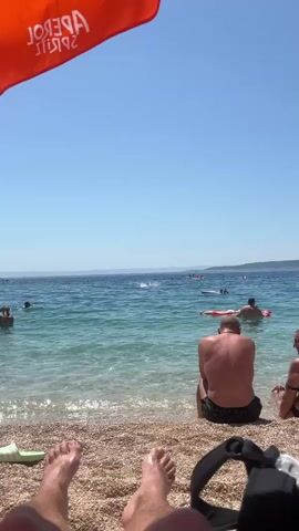 The video shows a beach scene. In the foreground, a person's feet are visible, resting on the pebble beach. To the right, a black backpack is placed on the sand. A beach umbrella, with the orange logo of "Aperol Spritz" visible, shades the upper left portion of the frame. The sea is calm and clear, with people swimming and relaxing on inflatable rafts. In the middle ground, a man is sitting on the beach facing the water. The horizon is visible in the distance, with landmasses or mountains. The sky is a clear blue. The overall impression is a sunny day at a crowded beach.