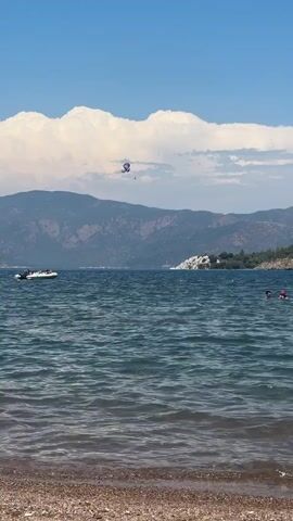 A scenic shot captures a bright, sunny day with fluffy white clouds dominating the sky. The scene is set at a tranquil beach with clear blue water lapping at the shore, which is covered in pebbles. In the distance, majestic mountains rise, their slopes covered in lush greenery.

The water is a hive of activity. A speedboat speeds across the surface, leaving a white wake behind it. Further out, two people are enjoying a swim, their heads visible above the waves. Dominating the sky is a parasailer, suspended high above the water, a colorful canopy against the blue canvas. The parasailer appears to be being towed by a boat, though the boat itself is not visible in the frame.

The overall mood is one of leisure and enjoyment, typical of a summer vacation. The clear skies and calm waters suggest pleasant weather, ideal for water sports and relaxation. The presence of the mountains adds to the picturesque quality of the location, hinting at a beautiful coastal landscape. The time of day is clearly daytime, given the brightness of the sun and the clear visibility. No visible text is present in the media.