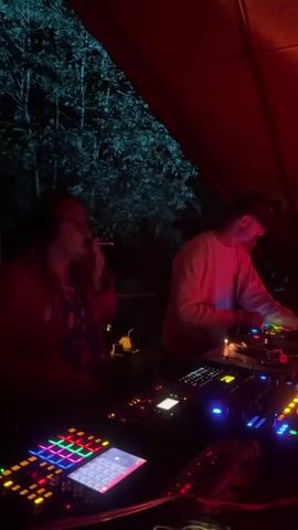 The video shows two men at a DJ booth outdoors at night under a red tent. The man on the left wears glasses and a red jacket over a patterned shirt, holding a cigarette and adjusting headphones. He operates a mixer with colorful illuminated buttons. The man on the right, wearing a white long-sleeved shirt and a dark baseball cap, is focused on the turntable and mixer in front of him. In the background, trees are visible, illuminated by blue-green light, and other people are partially seen. The DJ booth is equipped with multiple turntables, mixers, and controllers that light up in various colors.