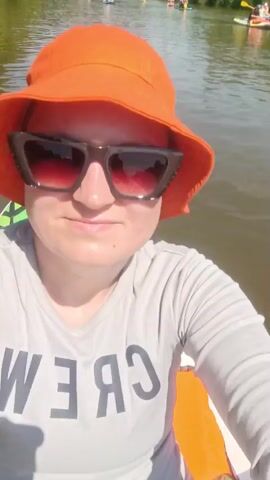 A woman wearing an orange bucket hat, sunglasses, and a gray long-sleeved shirt with the word "CREW" printed on it in black letters is paddleboarding on a river. She is looking directly at the camera and smiling slightly. Behind her, the river is visible, with several other people paddleboarding in the distance. The riverbanks are lined with green trees and vegetation. The sky is blue with some white clouds. The overall scene suggests a pleasant day for outdoor recreation.