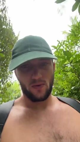 The video shows a shirtless man with a beard and a green cap walking through a lush, green forest. He is wearing a backpack and looking directly at the camera, smiling and saying, "Hey, do you know guys? I love you guys."  The scene is shot from a first-person perspective, likely by the man himself. The background is filled with dense foliage, suggesting he is hiking in a tropical or subtropical environment. The lighting is bright, likely daytime, with the sky visible through the canopy of trees.