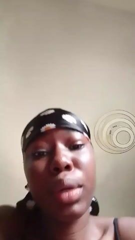 A close-up shot features a person with dark skin, wearing a black headscarf with white daisy patterns. They are looking directly at the camera and initially making a kissing face, then they smile. The person then touches their nose with their right index finger. The background is a plain, light-colored wall, with a decorative wire art piece. The subject wears a black camisole or tank top.