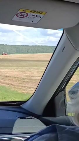 The video is taken from inside a car, looking out through the windshield. In the foreground, the car's dashboard and a part of the interior are visible. A sun visor with an airbag warning sticker is attached to the roof. Outside, a field is seen, which appears to be recently harvested, with rows of stubble. In the distance, a forest borders the field, and a red tractor is visible working in the field. A triangular road sign indicating a junction is also visible on the side of the road. The sky is partly cloudy with a contrail from an airplane.