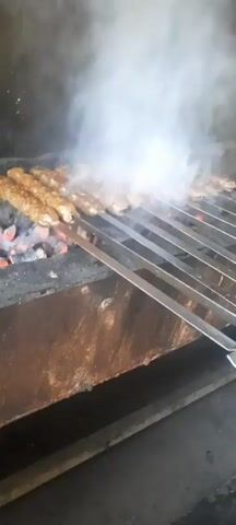 A close-up shot shows several rows of meat kebabs cooking over glowing red coals. The kebabs are skewered on long metal rods and arranged in parallel lines, with smoke rising from the cooking meat and coals. The scene appears to be in a kitchen or outdoor cooking area, with a rusty metal surface visible below the grill. The lighting is dim, highlighting the heat and intense cooking process.