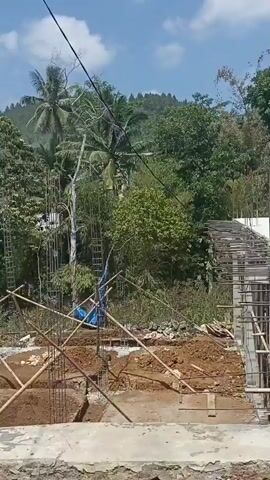 The video shows a construction site with visible foundations and rebar. Bamboo scaffolding is present, and a blue tarp is draped over some construction materials. The site is surrounded by lush greenery, including trees and palm trees, suggesting a tropical or subtropical environment. The sky is clear and blue. The camera pans across the site, showing different angles of the ongoing construction work. There are no people visible in the video.