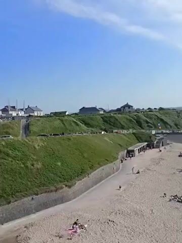 The video shows a wide shot of a sandy beach on a sunny day. The beach is populated with many people, some are sitting on towels, others are lying down, and a few are walking. There are also several blue tents scattered across the sand. In the background, there are green hills with some houses and a road. The ocean is visible, with some people in the water. The sky is blue with scattered clouds. The camera pans across the beach, providing a panoramic view of the scene.