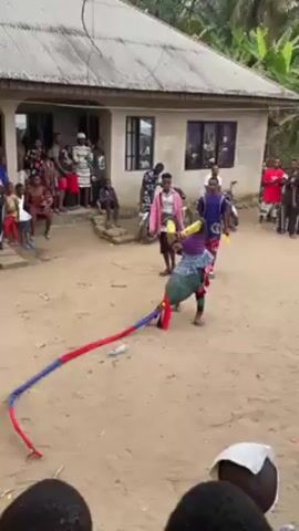 The video depicts a cultural performance taking place in an outdoor setting, likely a village or rural area, in front of a small house with a corrugated iron roof. A performer, dressed in a colorful costume and wearing a headdress, dances while holding two long, ribbon-like streamers, one red and one blue. They move in a crouched, energetic manner, twirling the streamers. A small crowd of people surrounds the performance area, with some watching from the porch of the house and others standing around the perimeter. Many are casually dressed. A few individuals are observing the performance while standing near a motorcycle parked in the background. The ground is sandy, and some trees and foliage can be seen in the background. The overall atmosphere appears to be lively and celebratory.