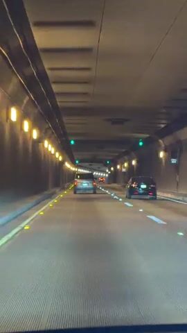 This is a first-person perspective video traveling through a tunnel. The tunnel is lit by rows of yellow lights along the walls and green traffic lights overhead. Several cars are visible on the road ahead, all traveling in the same direction. The road surface appears to be made of grooved concrete. The video is shot from inside a vehicle, looking forward.