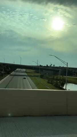 From a car, the view shows the sun shining through clouds in a light blue sky. Palm trees line a green space with a pond next to a highway. A concrete median separates the lanes. Cars are visible on both highways. Above, highway overpasses are seen.