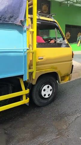 The video begins with a close-up shot of a light blue truck bed with a large blue tarp covering its contents. Yellow metal bars are visible along the side of the truck. The camera then pans to the rear of the truck, revealing its taillights and license plate. The truck is parked on a wet, asphalt surface, suggesting recent rain or cleaning. As the video progresses, the truck begins to move forward. A woman on a motorcycle, wearing a dark headscarf and a light-colored shirt, passes by the truck on the right side of the road. The surrounding environment appears to be a street or alleyway with buildings on either side. The buildings have colorful exteriors, including a prominent green structure. Utility poles and wires are also visible in the background. The overall scene depicts an outdoor environment, possibly in a residential or commercial area, with the truck and motorcycle being the primary subjects of action.