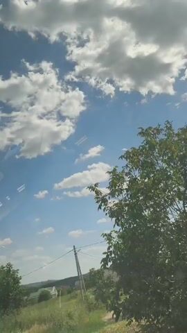 A time-lapse video shows a bright blue sky filled with white, fluffy clouds. The camera angle is tilted upwards, suggesting it was filmed from a vehicle moving through a rural or suburban area. On the left side of the frame, a triangular road sign with red borders and a white background depicts two arrows pointing upwards, indicating a two-way traffic situation. Green trees and vegetation are visible along the right side of the frame. In the distance, houses and buildings can be seen amidst rolling hills. A dirt road is also visible on the right. A utility pole with wires stretches across the middle of the frame. The overall impression is one of a sunny, daytime scene with passing clouds.