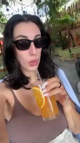 A woman with black hair and large sunglasses is drinking a beverage with a straw. The drink is in a clear glass and contains a slice of orange. She is wearing a brown tank top and a light blue shirt draped over her shoulder. The setting appears to be an outdoor cafe or restaurant with greenery and other seating visible in the background. The woman smiles at the end of the video.