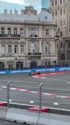 The video shows a Formula 1 race in an urban environment. The camera is positioned behind a metal barrier, looking across the track. An orange race car with visible sponsor logos passes quickly from right to left. Behind the track are barriers with "AMERICAN EXPRESS" written on them. In the background, there are tall, ornately decorated buildings with domed roofs. A large screen displays live footage of the race. People can be seen on the other side of the track, near the barriers. The sky is overcast.