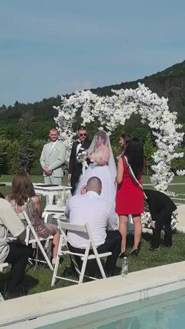 An outdoor wedding ceremony takes place on a sunny day on a green lawn with a white floral arch in the background. A bride in a white gown and veil stands beside a groom in a black tuxedo and sunglasses. They are holding hands. To their left, a man in a light green suit stands. To their right, a woman in a red dress with a green sash stands. Several guests are seated on white chairs in the foreground, facing the couple. A man in a white shirt has his back to the camera. A child in a black suit is bending down on the far right. A swimming pool edge is visible in the extreme foreground. Green trees and hills are in the distant background.