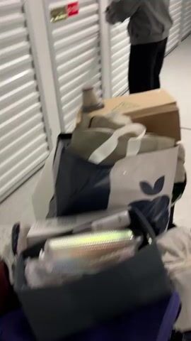 A woman opens the door to a storage unit in a corridor with rows of units. She wears a grey bomber jacket and black pants.  A dolly loaded with items sits in front of the unit. The dolly holds a cardboard box, a blue and white shopping bag, and a grey storage box containing a shiny water bottle. The storage unit door is grey with a red label reading "01-7258."  The corridor is brightly lit with a grey floor and white doors. The woman uses a key to open the unit.