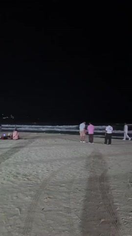 The video shows a nighttime scene at a beach with several people present. The sand is visible with tire tracks. Waves are breaking on the shore, and the sky is dark. In the foreground, there are tire tracks on the sand. Further back, a group of four people are standing near the water, with one person wearing a pink shirt. To the right, a red flag is planted on the beach. The camera then pans to the left, showing a group of people sitting on the sand in the distance. Finally, the camera pans to the right again, showing more people walking and sitting on the beach. Lights from buildings or a town are visible in the far distance along the coastline.