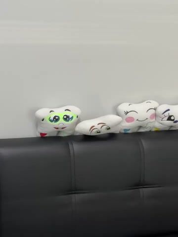 A black leather couch is against a white wall. On the couch are several plush toys. Along the back of the couch are small plush teeth with cartoon faces. There are at least eight of these. To the right and closer to the viewer are larger plush toys. One is a green creature with a large mouth full of teeth and red accents. Another is a pink cat-like creature with a similar large mouth full of teeth. There is also a plush lion and a long pink stuffed object that resembles a tongue. A yellow toothbrush is visible.