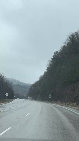 The video shows a car driving on a wet, two-lane road, possibly in a mountainous area. The sky is overcast and gray, indicating rainy or cloudy weather. On either side of the road, there are trees, with a dense forest visible on the right side of the road rising up a steep hill. In the distance, there are more hills partially obscured by mist or fog, with what appears to be a light dusting of snow on the tree branches. There are three cars visible on the road, moving in the same direction as the camera car. There are road signs on both sides of the road, though their details are not clear. The road surface is wet, reflecting the gray sky, and the camera appears to be mounted inside a car, looking out through the windshield.