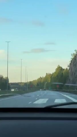 The video shows a car driving on a highway during daytime. The sky is blue with some light clouds. The highway has multiple lanes and is surrounded by trees and a rock face. There are other cars on the road, and streetlights line the highway. A speed limit sign is visible on an overpass. The view is from inside the moving car, through the windshield.