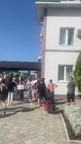 A group of people are standing outside a white building with a green roof, holding luggage. The building has multiple windows and air conditioning units. There is a large green bush in the foreground. One person in the foreground is wearing a white cap and carrying a backpack. In the background, a banner is visible on the building that reads "ЖЕМЧУЖИНА РОССИИ" which translates to "Pearl of Russia." The sky is clear and blue. The people appear to be arriving or departing from the building. Some people are looking at their phones, while others are talking to each other. The overall impression is of a sunny day at a hotel or resort.