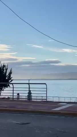 The video presents a harbor scene. The camera pans from left to right to reveal a railing along the waterfront, with distant mountains visible across the water. A cruise ship is docked at the harbor. The sky is blue with scattered clouds, and the water is calm. A street with red and yellow markings is in the foreground. A person is visible sitting on the edge of the waterfront. There are lampposts and a small structure with the text "Cumplimos" in the harbor area.
